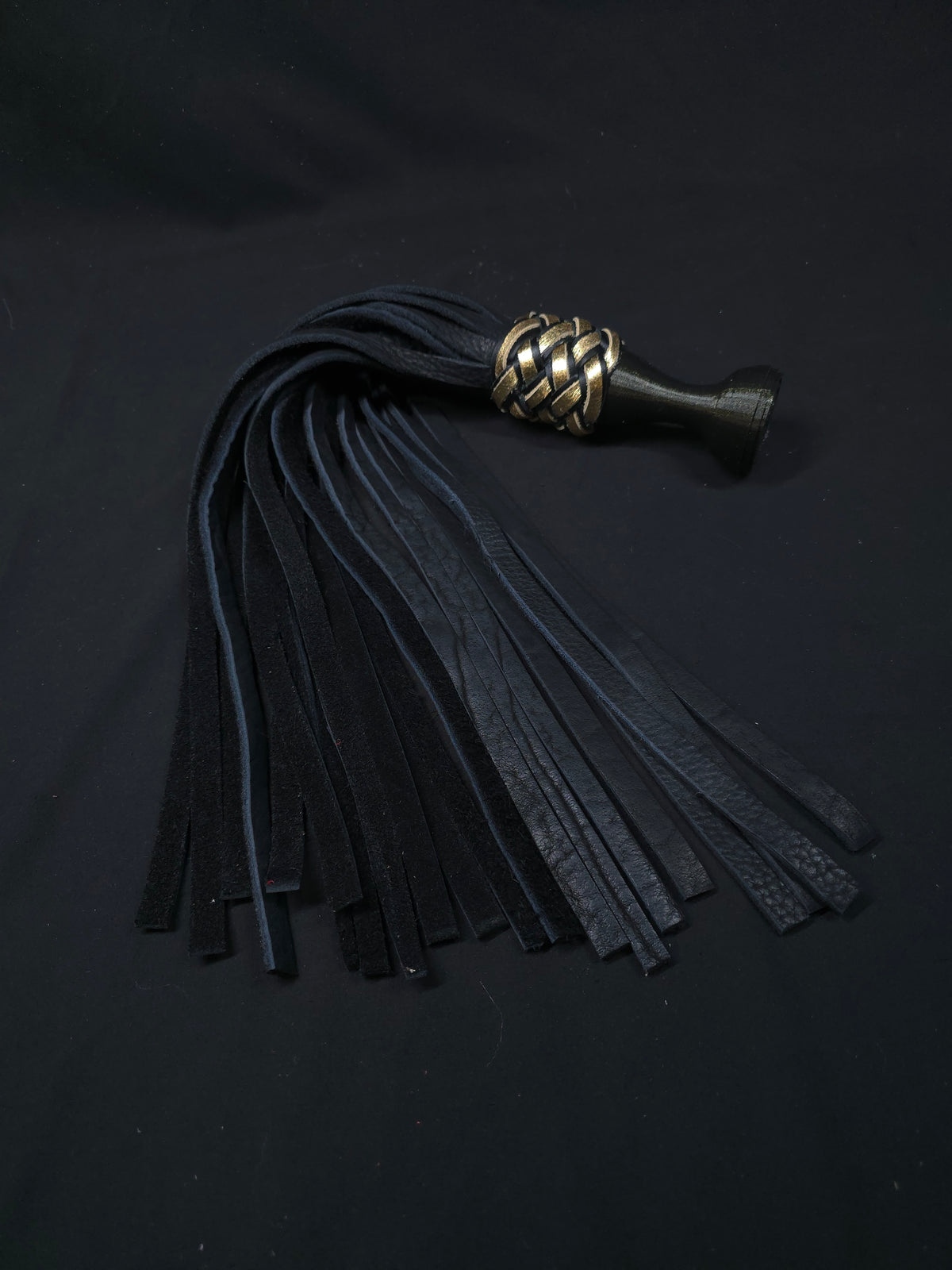 Black leather flogger with a braided gold accent and a ball handle meant to sit between the index and middle finger with the flat top pressed against the palm. There is a triskellion engraved on the top of the handle. Displayed on a black fabric background.